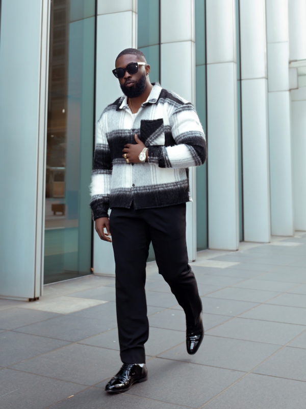 A Man In A Black And White Patterned Jacket And Sunglasses Walking Along A Modern Glass Walled Building.